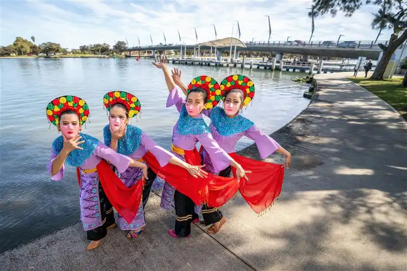 Four performers in colourful traditional outfits pose by the water, holding red cloths as part of their dance.